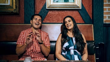 Dean Obeidallah and Maysoon Zayid sitting side-by-side on a wooden bench laughing and smiling.