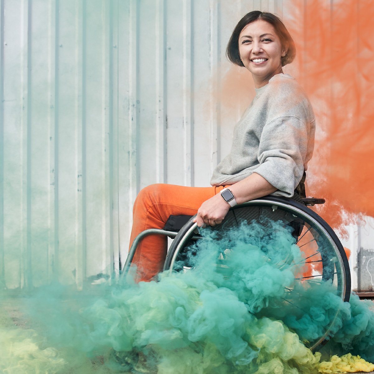 woman in wheelchair smiles to camera with orange and green smoke flares blossoming around her.
