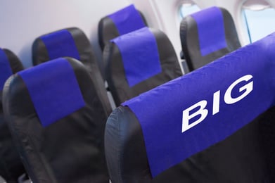 A row of airplane seats with blue covers over the headrests.  One seat's headrest cover says "BIG" indicating the seat is designed for plus-size passengers