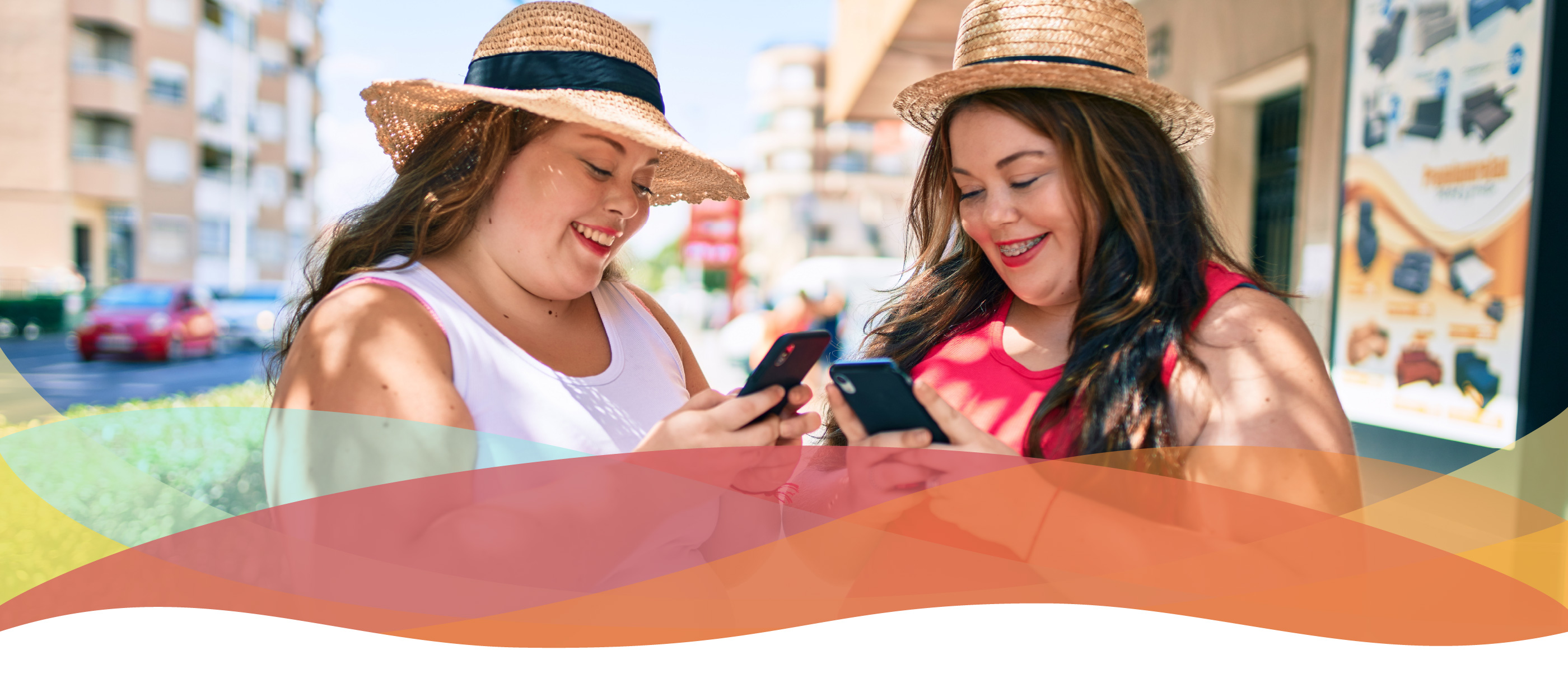 Two plus-sized twin sisters check their phones, outside.  Both are wearing straw hats, tank tops, white jeans, and big smiles.