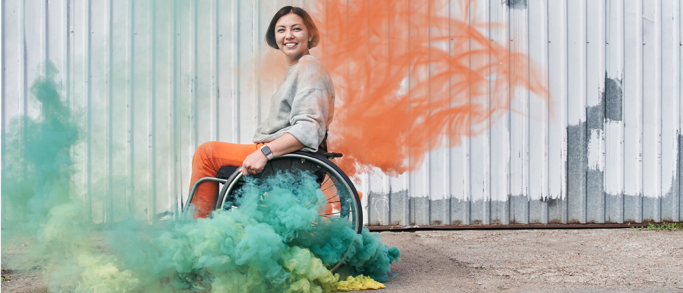 woman in wheelchair smiles to camera with orange and green smoke flares blossoming around her.