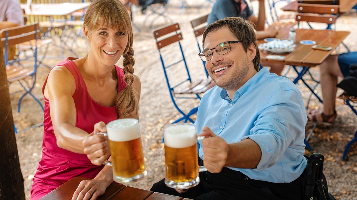 A man in a wheelchair dines at a table with a friend, raising beer in toast.