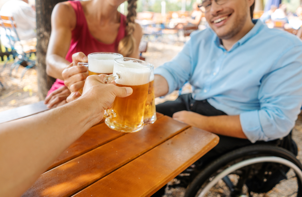 A man in a wheelchair dines at a table with a friend, raising beer in toast.