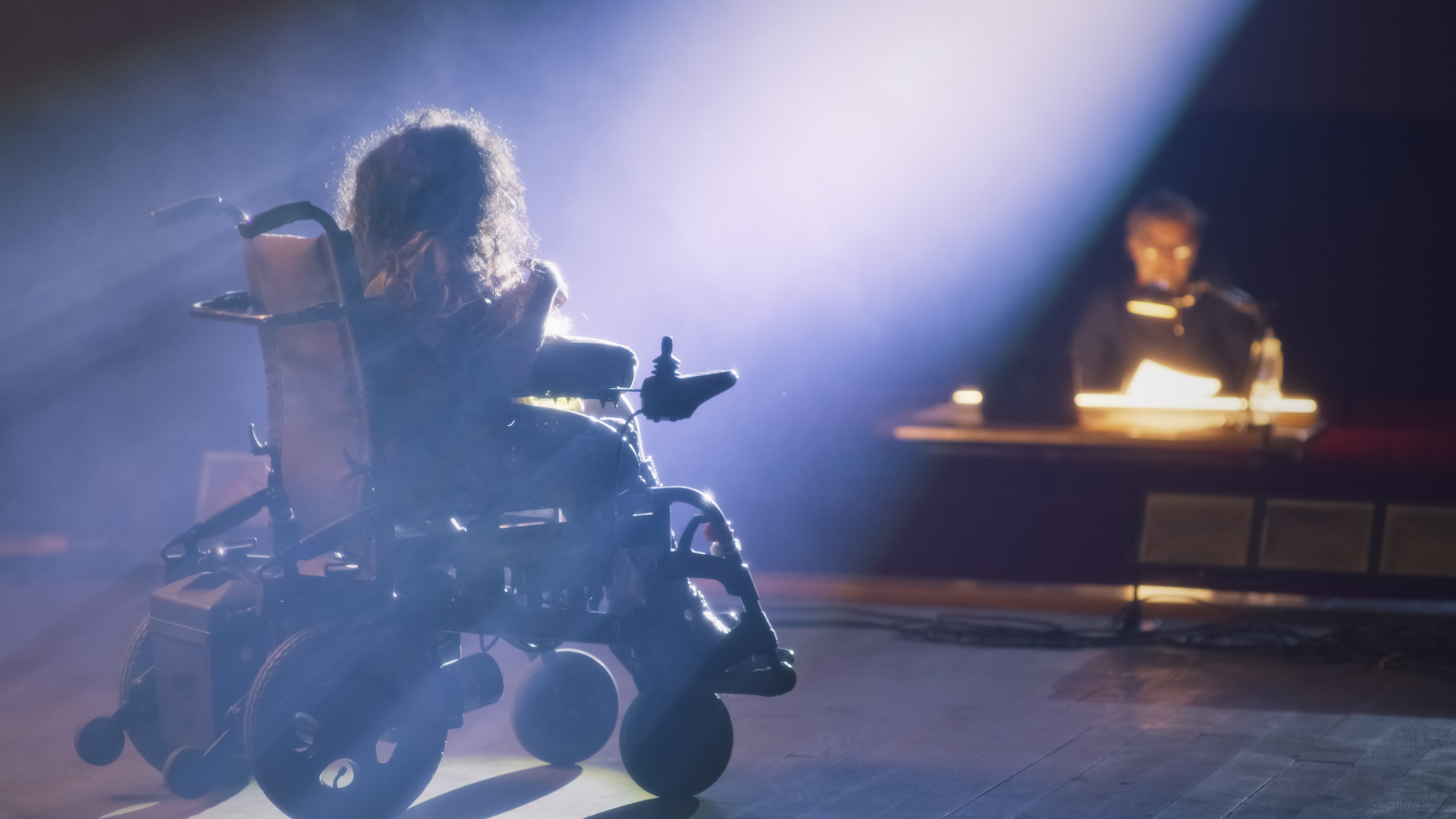 Actress with a disability driving around the stage in an electric wheelchair and rehearsing a performance with actors and a director in a dim theater, with a spotlight