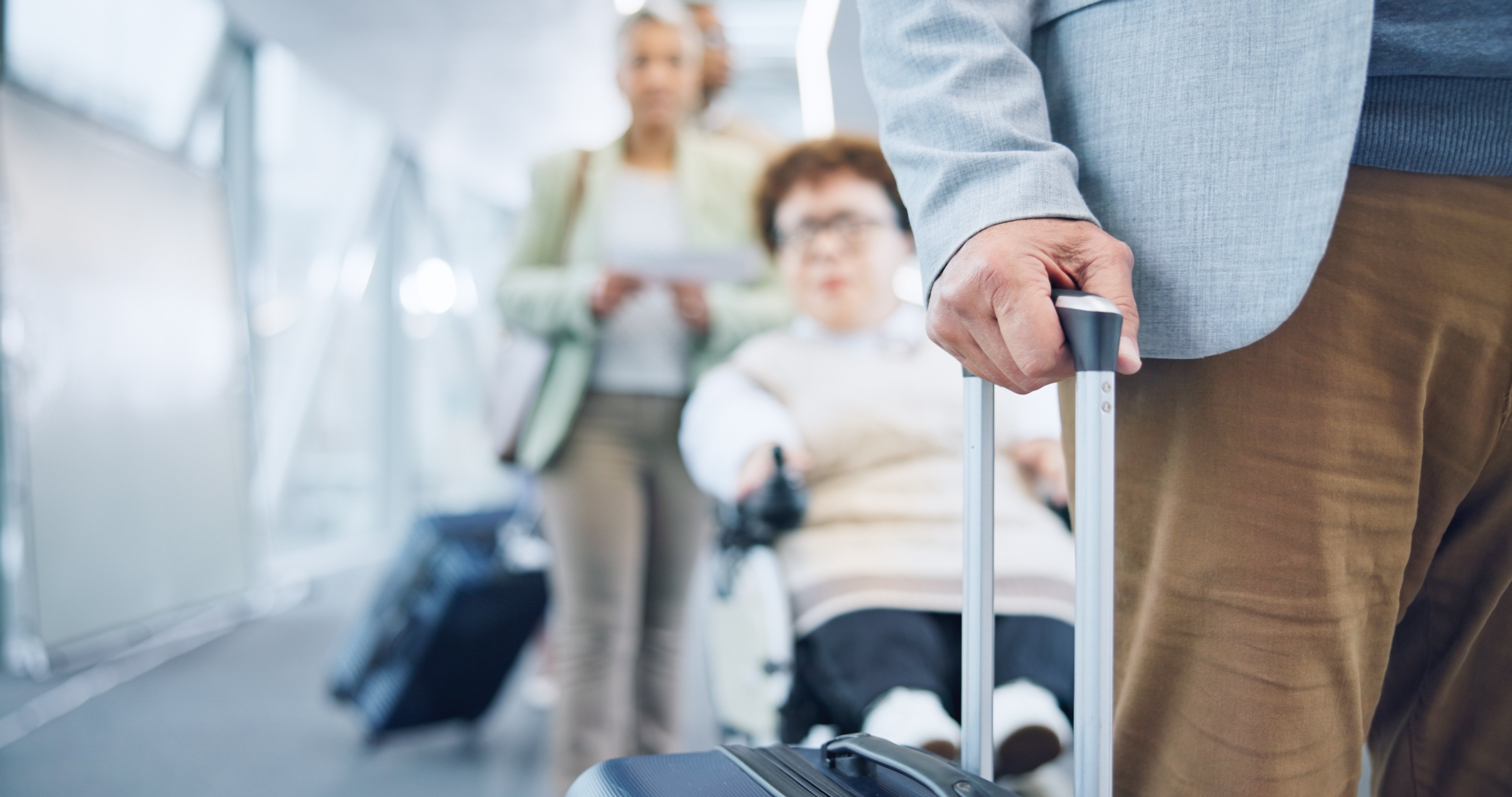 A close-up image of a hand holding the handle of rolling luggage, with a blurry background which has a wheelchair user in line behind them.